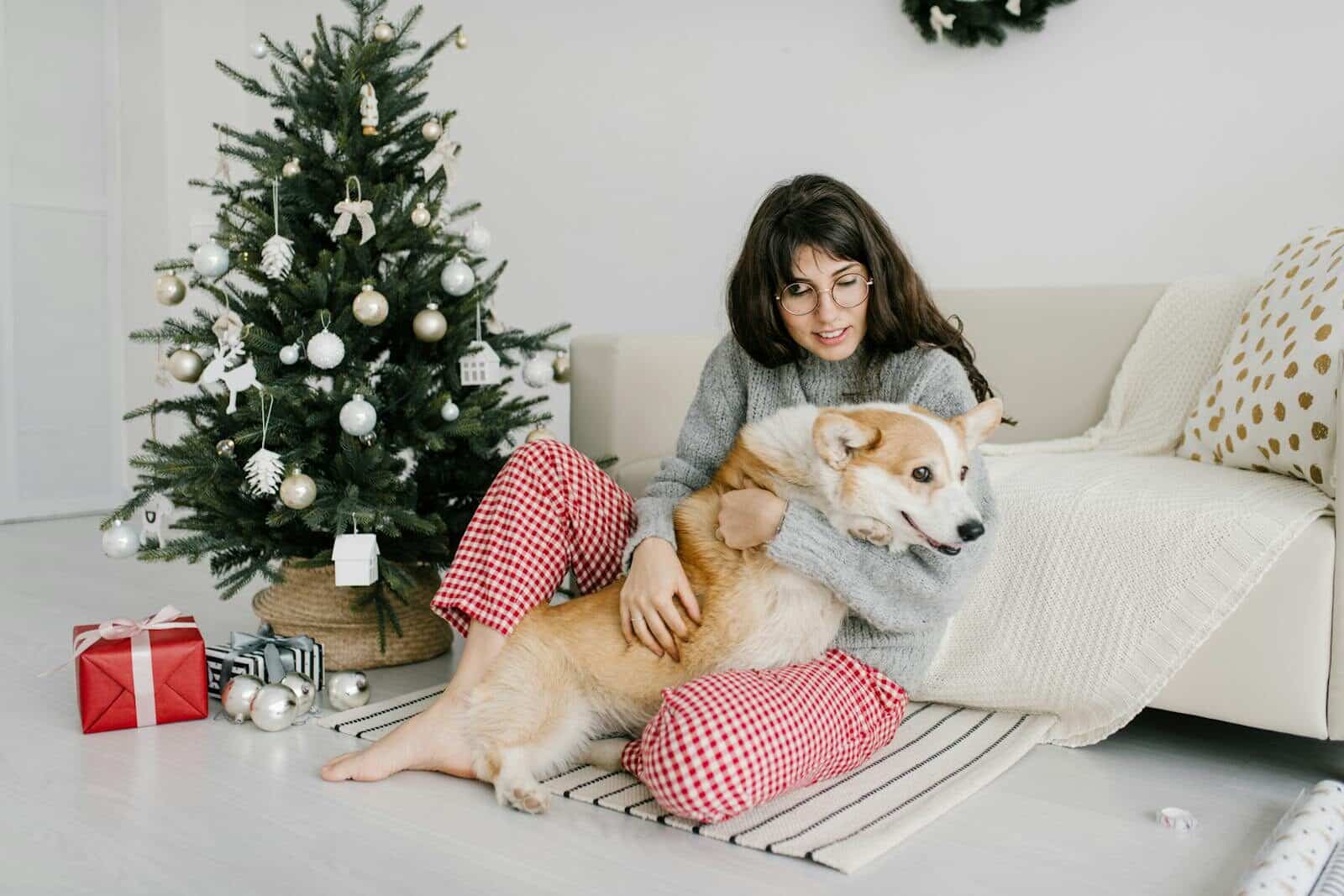 Photo by Julia Volk A woman sits by a Christmas tree with her dog, enjoying the festive atmosphere.