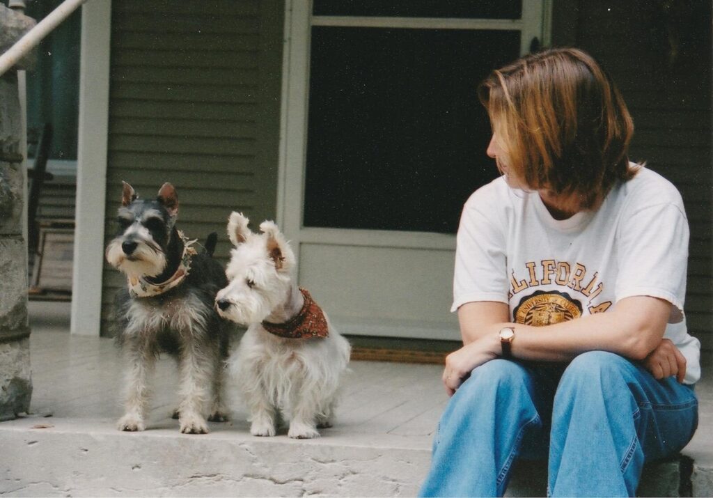 A woman sits with two Schnauzer dogs on a porch, capturing a serene and domestic moment.
